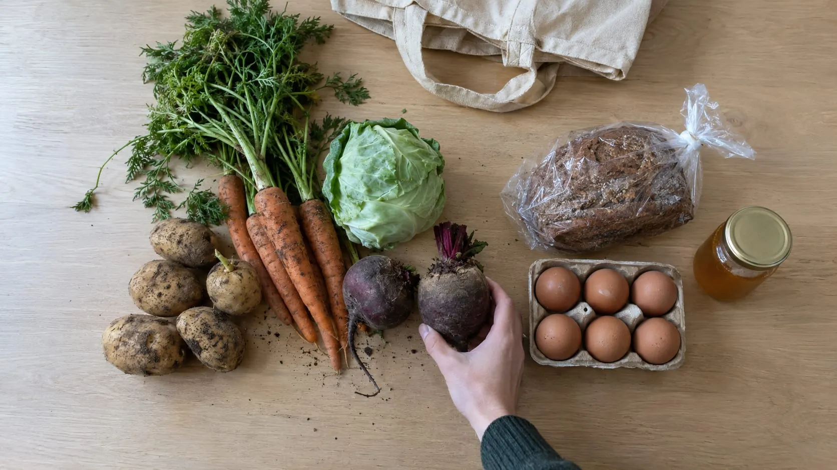 Pöydällä multaisia porkkanoita, perunoita, kaalinpää, punajuuria, ruisleipää, kananmunia ja hunajapurkki sekä kangaskassi